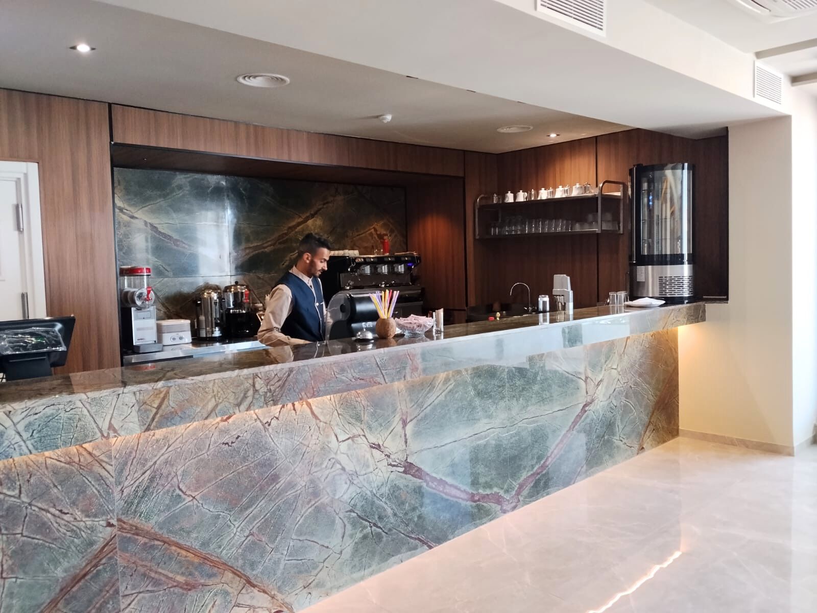 Un hall d’hôtel moderne avec un comptoir d’accueil en marbre, une machine à café et un grand écran fixé au mur.