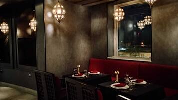 A dimly lit restaurant with tables set for dining, a red cushioned bench, and hanging light fixtures.
