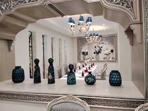 Un comedor con una mesa blanca dispuesta para una comida, rodeado de decoraciones ornamentales en la pared y un espejo grande.