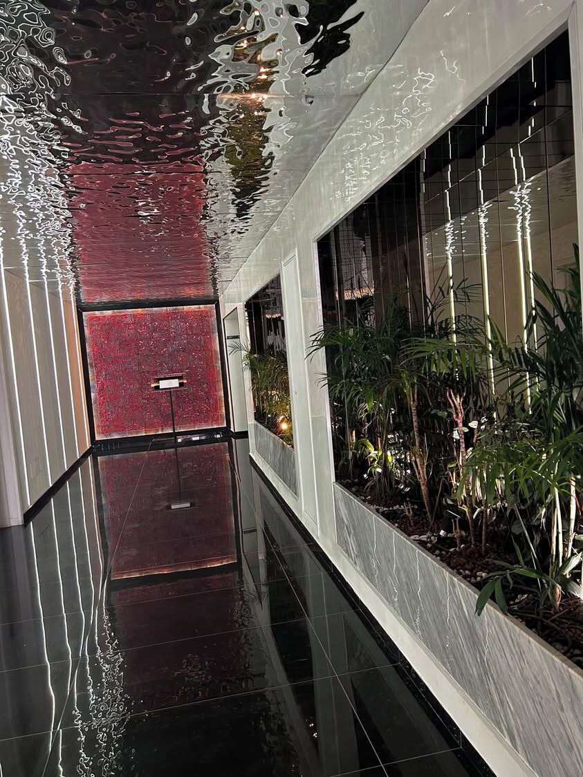 Un pasillo con piso reflectante, pared texturizada de color rojo y una jardinera con plantas verdes.