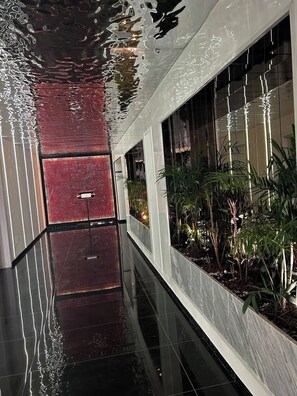 Un pasillo con piso reflectante, pared texturizada de color rojo y una jardinera con plantas verdes.