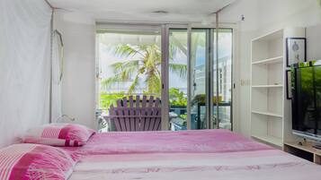 A bedroom with a bed, a TV, a balcony with a chair, and a view of palm trees.
