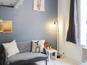 A modern living room with a grey sofa, a small white table with decorative items, a floor lamp, and a window with curtains. - Studio Mezzanine – Near Metro Line 13 (Saint-Ouen-sur-Seine)