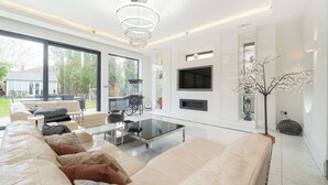 A modern living room with a large sofa, a glass coffee table, and a flat-screen TV mounted on the wall. - Spacious 5-Bed Family Home with Garden & Parking (London)
