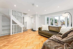 A modern living room with a herringbone wooden floor, a sofa with cushions, a staircase, and large windows. - Designer 4-Bed, Skyline Views & Free Parking (London)