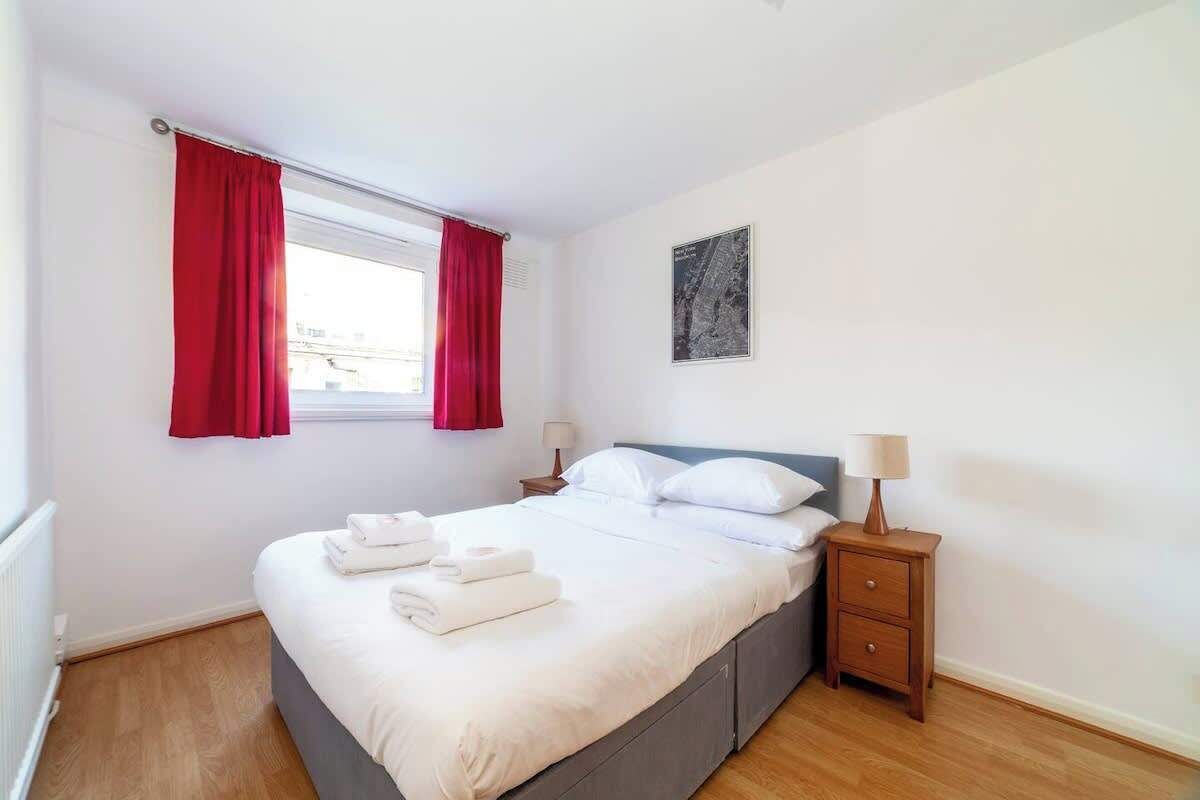 Cama bem arrumada com lençóis brancos, uma pequena mesa de cabeceira com abajur, uma janela com cortinas vermelhas e um mapa fixado na parede.