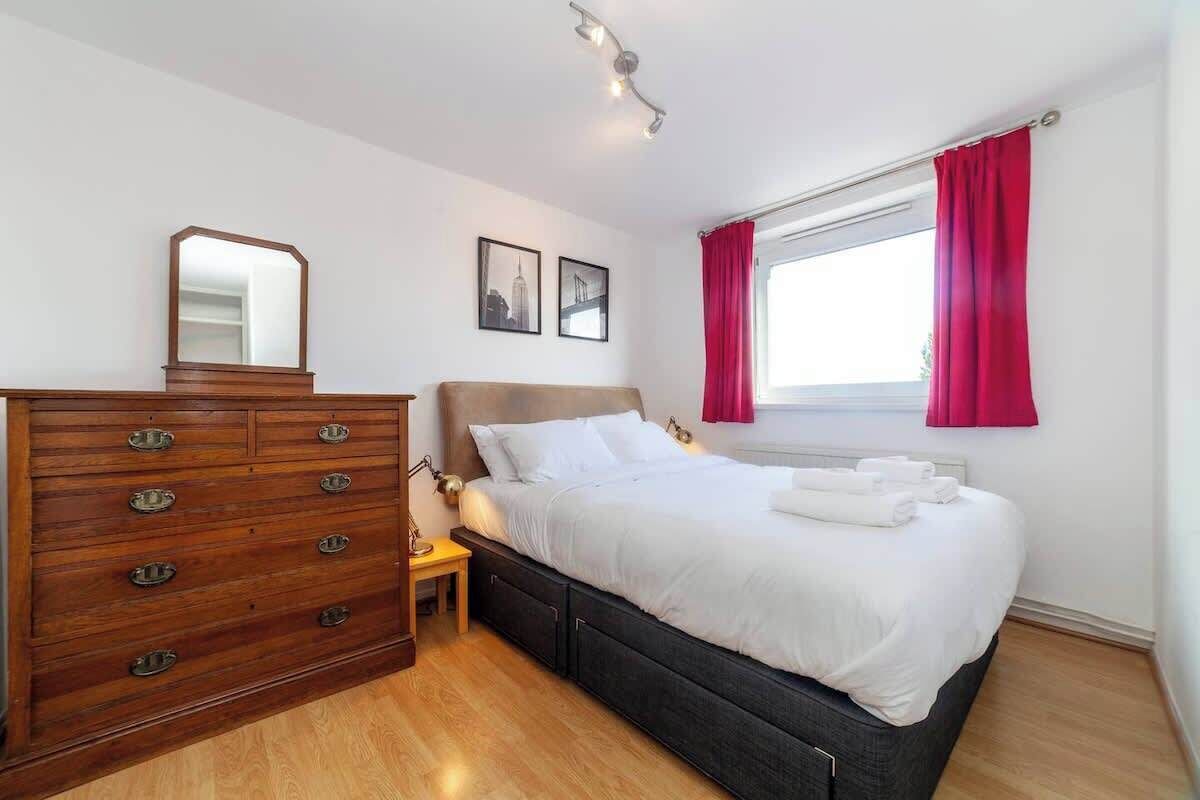 Une chambre avec une commode en bois, un lit recouvert de draps blancs, une table de chevet, un miroir et deux tableaux encadrés accrochés au mur.