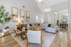 Ein geräumiges Wohnzimmer mit grauem Sofa, weißen Sessel, Couchtisch und Essbereich mit Holzsesseln und Tisch.
