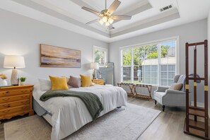 Ein Schlafzimmer mit einem Bett, Nachttischen, einem Sessel, einem Deckenventilator und großen Fenstern.
