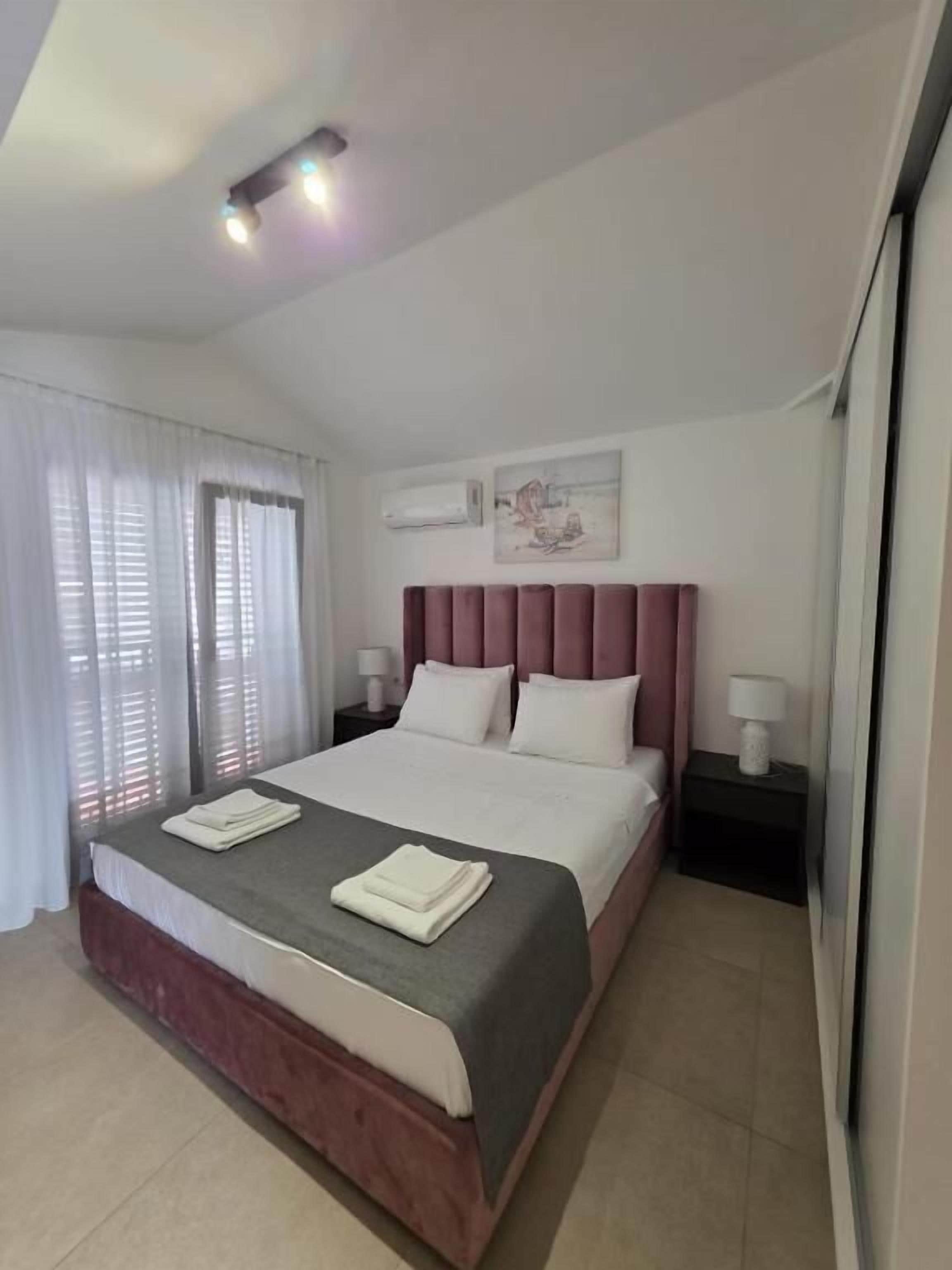 Un dormitorio con cama, mesitas de noche, una ventana con cortinas e iluminación en el techo.