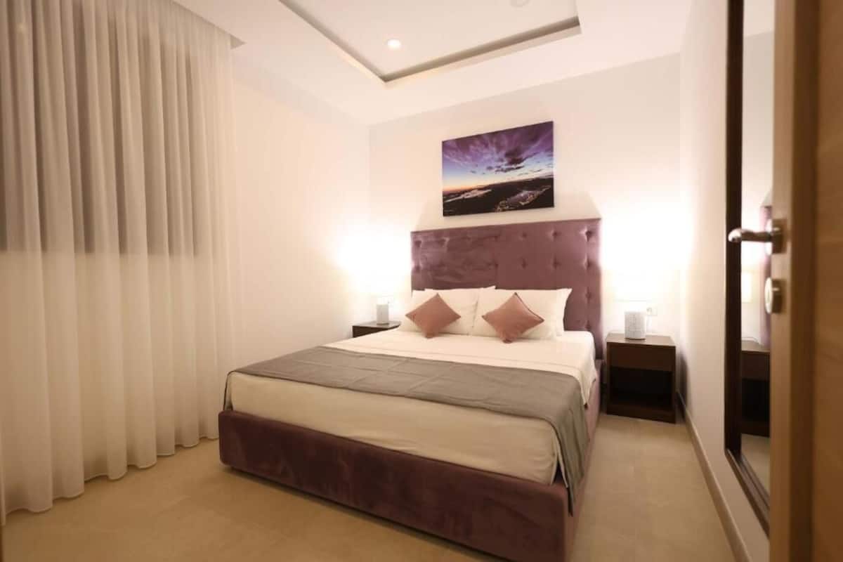 A hotel room with a large bed, a headboard with a cityscape painting, two bedside tables with lamps, and a large window with curtains.