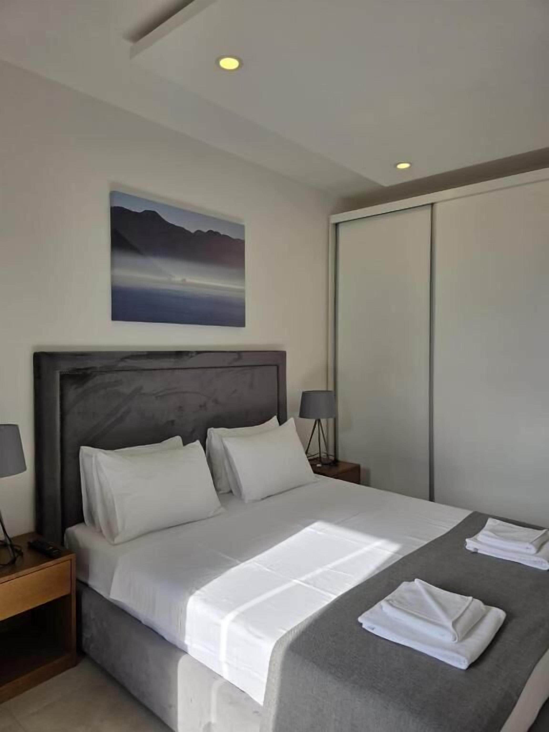 A neatly made bed with white linens, a headboard, bedside lamps, and a painting above the bed.