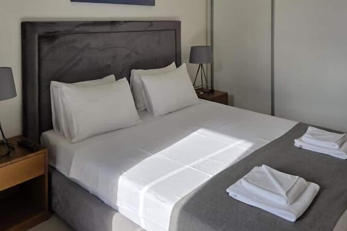 A neatly made bed with white linens, a headboard, bedside lamps, and a painting above the bed.