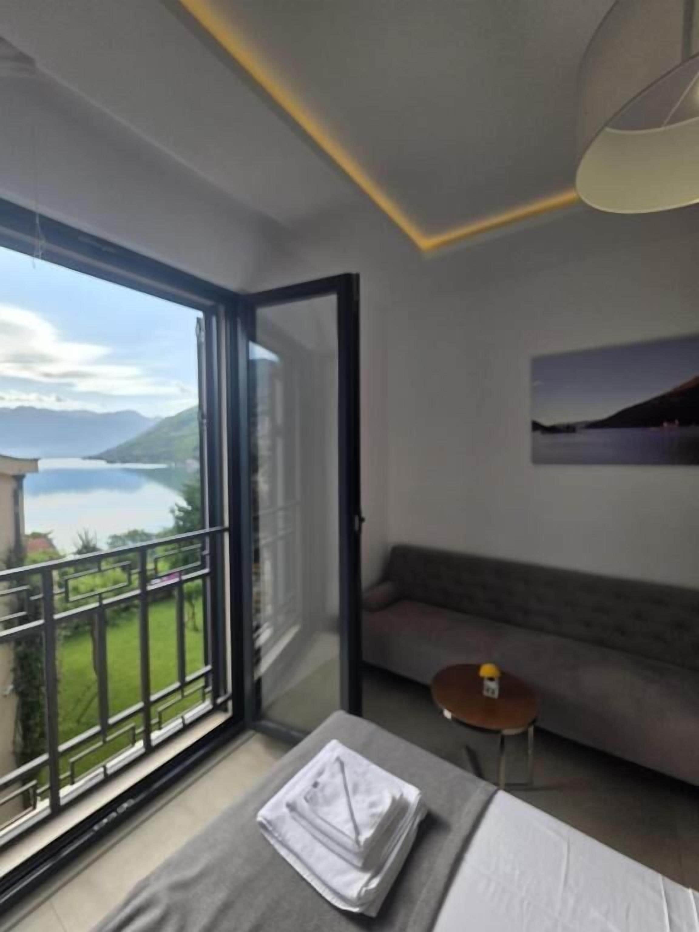 A hotel room with a large sliding glass door leading to a balcony with a view of a lake and mountains. There is a bed with a neatly folded white towel and a small round table with a bottle.