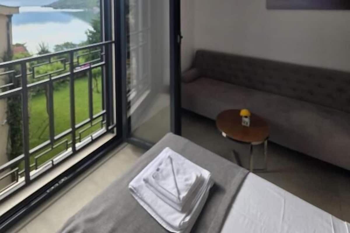 A hotel room with a large sliding glass door leading to a balcony with a view of a lake and mountains. There is a bed with a neatly folded white towel and a small round table with a bottle.