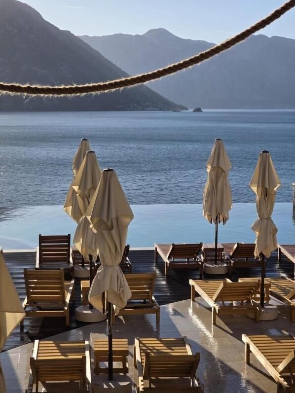 A poolside area with wooden lounge chairs and umbrellas, overlooking a calm body of water and mountains.