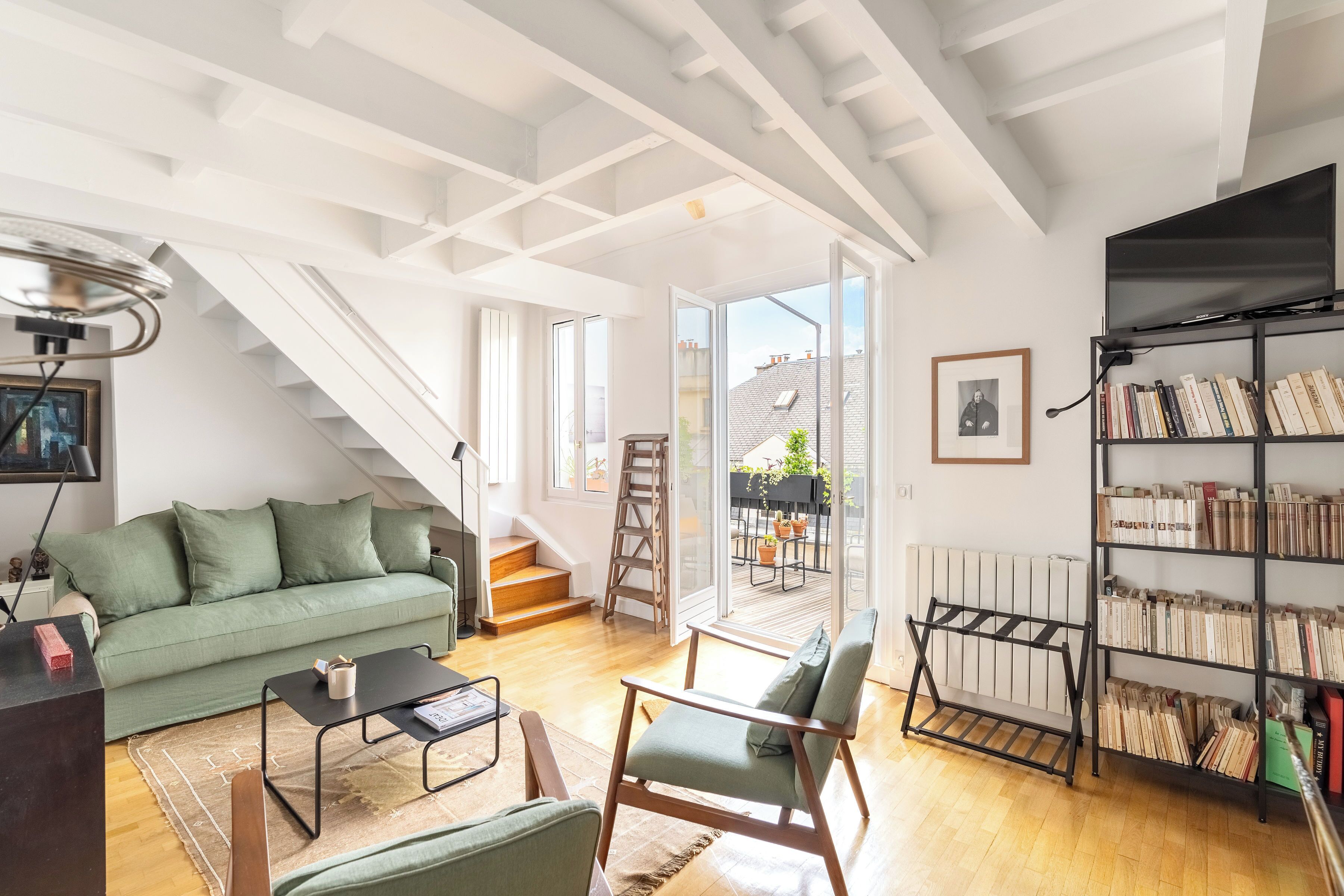 A modern living room with a green sofa, a coffee table, a bookshelf, and a staircase leading to an outdoor area.