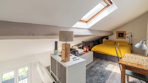 Et soveværelse med skunk og ovenlys, en seng med gul sengetøj, et skrivebord med lampe og et lille bord med flettede underlag.