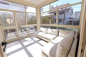 A sunlit room with a beige sofa, large windows, and a view of buildings outside.