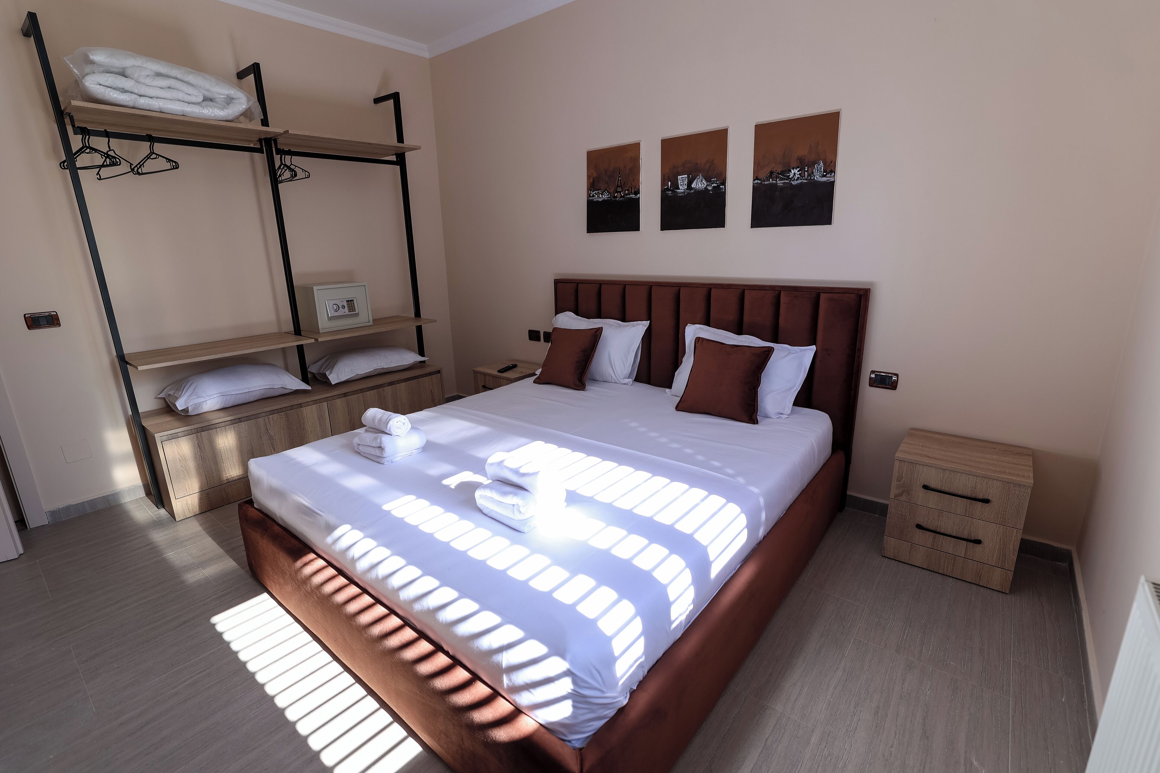 Una habitación de hotel ordenada con una cama grande, una mesita de noche, una repisa en la pared con toallas y tres obras de arte.