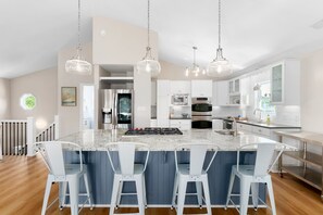 A modern kitchen with a central island, white bar stools, stainless steel appliances, and pendant lighting. - Second Wynd, Luxury Home, Private Pool, Elevator, NewlyUpdated (Emerald Isle)