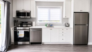 Una cocina moderna con electrodomésticos de acero inoxidable, armarios blancos, un ventanal con persianas y suelo de madera.