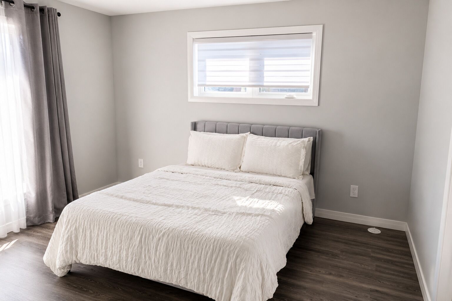 Une chambre à coucher avec un lit, des rideaux gris, une fenêtre et une tête de lit blanche.