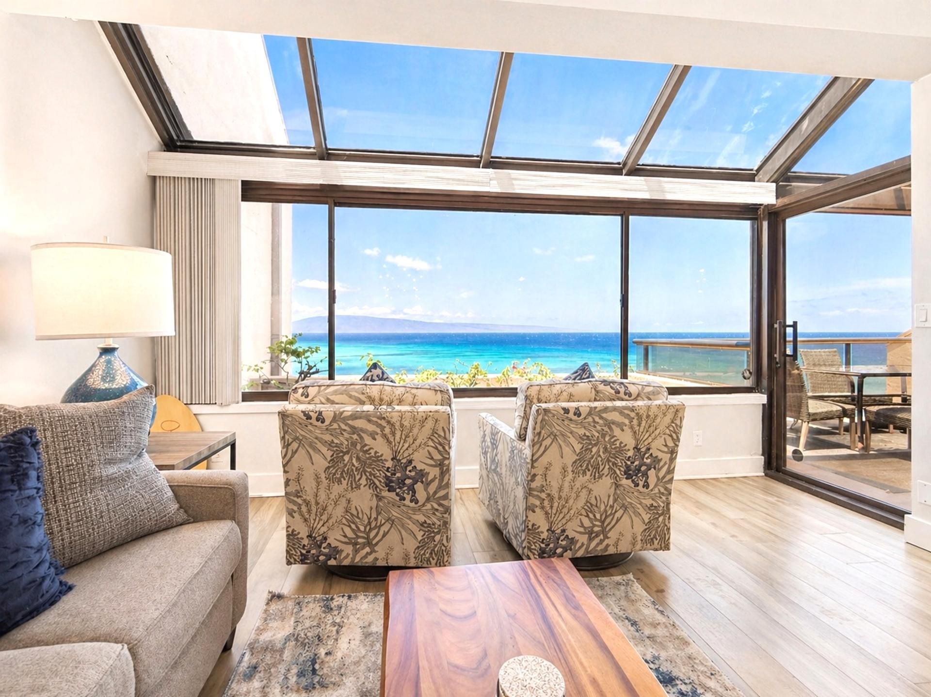 Un salon doté d’une grande baie vitrée donnant sur une plage. On y trouve un canapé, deux fauteuils et une table basse.