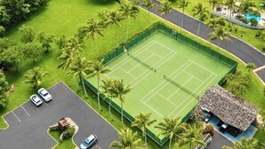 An aerial view of a tennis court surrounded by palm trees, with a parking area and a building with a thatched roof. - Kuilima Estates 2 Golf View Villas, Sleeps 16 | Car Incl. w/ 6 Nights | KUL ML-4046 by KBM (Kahuku)