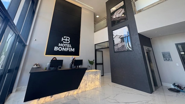 A modern hotel lobby with a reception desk, a large black sign with the hotel name, and a staircase leading to the upper floor. - Hotel Bonfim (Silvânia)