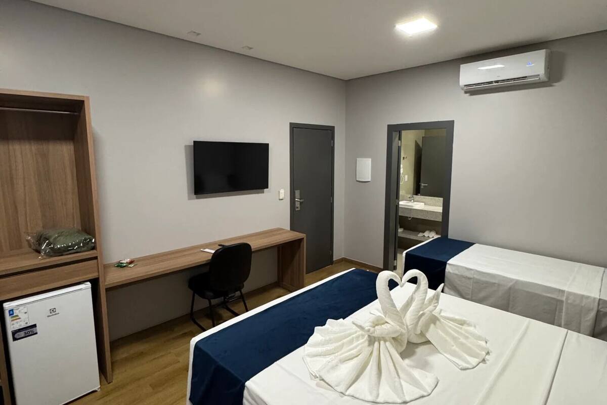 Habitación de hotel con dos camas, un escritorio, una silla, un refrigerador pequeño y un equipo de aire acondicionado.
