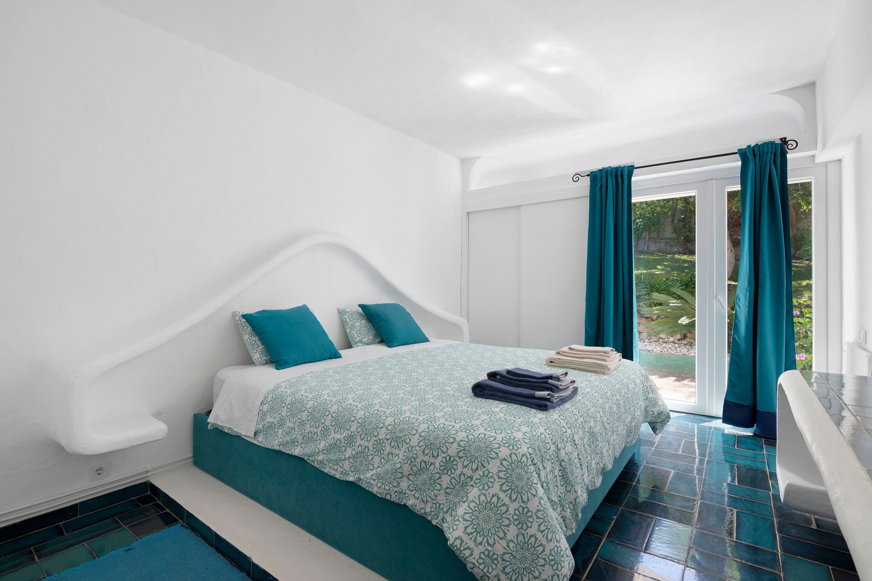 A bedroom with a bed, teal curtains, a white wall, and a view of greenery outside.