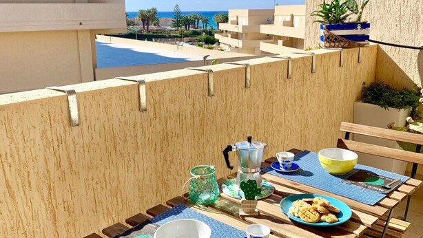 A balcony with a table set for breakfast, a view of the sea, and a building with balconies. - La terrazza sul blu - Marina di Ragusa (Marina di Ragusa)