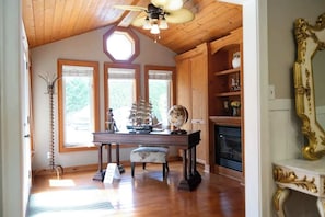 A room with wooden flooring, a ceiling fan, a table with a globe and ship model, a fireplace, and a large ornate mirror.