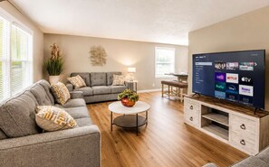 A modern living room with a grey sofa, a round coffee table, a TV on a wooden stand, and a potted plant. - Hot Tub • Game Room • 5BR • Suburb near Everything (Rochester)