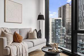 Ein modernes Wohnzimmer mit grauem Sofa, einer schwarzen Stehlampe, einem runden Couchtisch und einem großen Fenster mit Blick auf die Stadt.