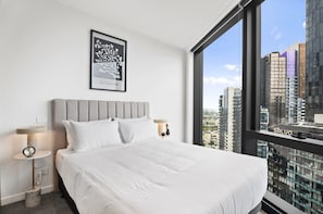 Ein Hotelzimmer mit Bett, einem gerahmten Kunstwerk, einem Nachttisch, einer Lampe und Blick auf eine Stadtlandschaft durch große Fenster.