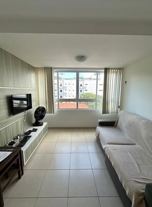 Ein Wohnzimmer mit einer Couch, einem an der Wand befestigten Fernseher, einem Ventilator und einem Fenster mit Blick auf Gebäude.