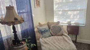 Una habitación con suelo de madera, un sillón redondo con una manta suave, una lámpara, una planta en maceta y una ventana con persianas.