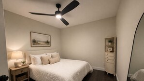 A bedroom with a bed, nightstand, lamp, ceiling fan, and a small shelf with a plant. - Charming 2BR Guest House w/ Pool - Perfect Getaway (West Tisbury)