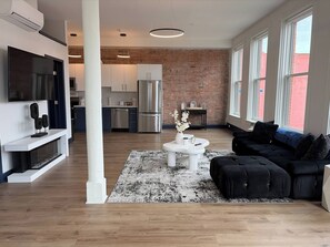 A modern living room with a black sofa, a white coffee table, and a TV on a white stand. - Bright Sunlit Studio in Downtown Rochester Floor-to-Ceiling Windows (Rochester)