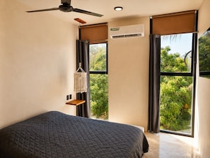 A bedroom with a bed, a ceiling fan, an air conditioning unit, a macrame wall hanging, and large windows with a view of greenery. - Modern Flat in Zicatela • 100m from the Beach (Oax.)