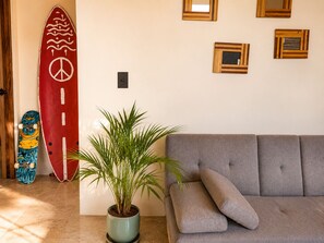 A room with a grey sofa, a potted plant, a surfboard, and wooden wall decorations. - Modern Flat in Zicatela • 100m from the Beach (Oax.)