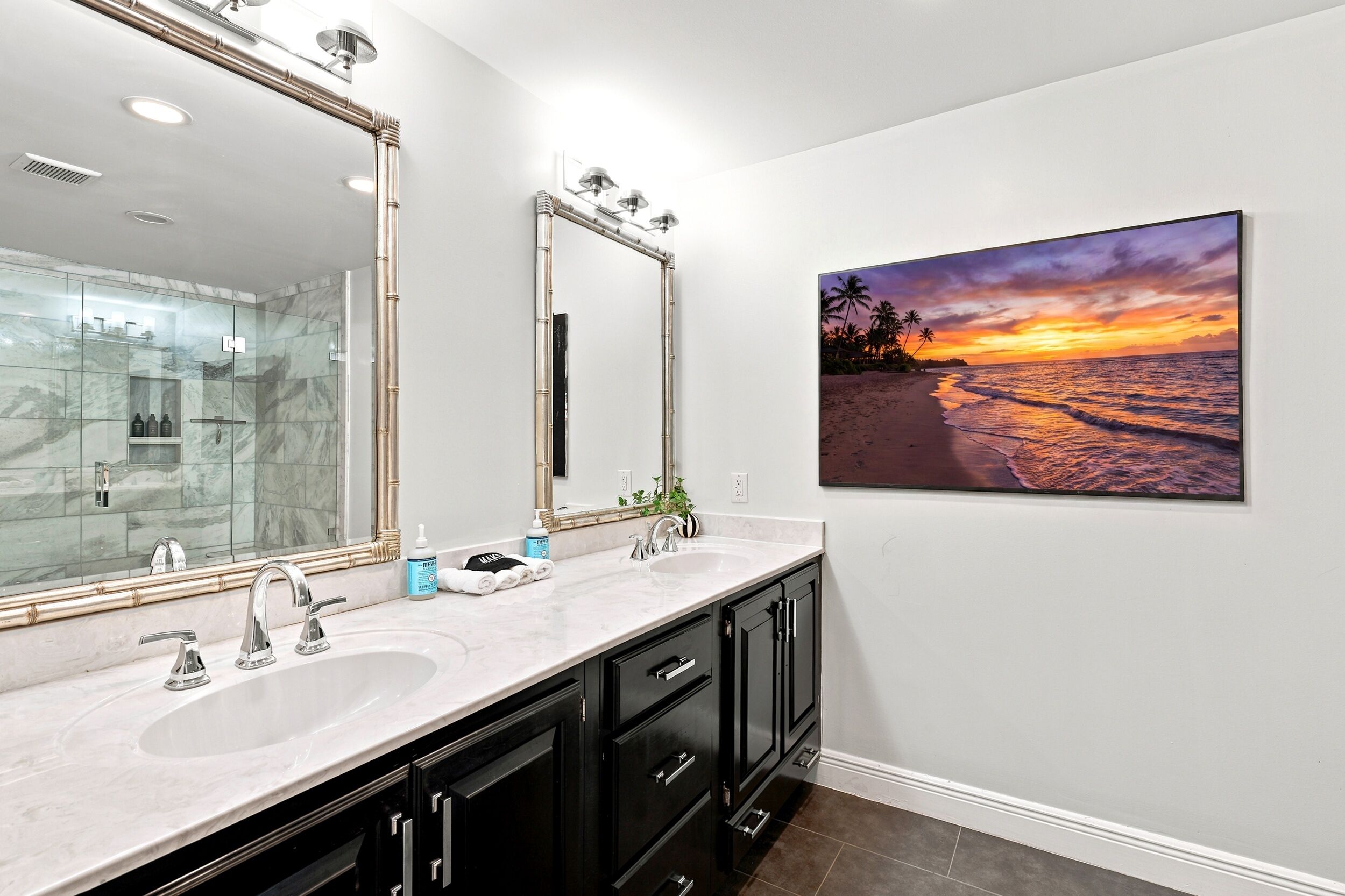 Une salle de bain avec un grand miroir, deux lavabos et un tableau représentant un coucher de soleil sur une plage.