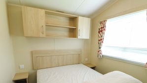 A bedroom with a bed, a headboard, a wardrobe, a window with curtains, and a small bedside table. - J J Caravan (Abergele)