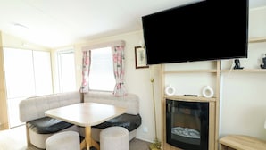 A living room with a sofa, a table, ottomans, a flat-screen TV, a fireplace, and a window with curtains. - J J Caravan (Abergele)