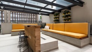 A modern interior with a glass ceiling, a wooden reception desk, a yellow sofa, and a bar area. - Uwin 403 - Apartment 10 minutes from Chucri Zaidan (São Paulo)