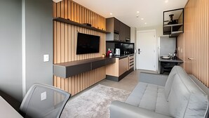 A modern living room with a grey sofa, a flat-screen TV mounted on a wooden panel wall, a kitchen area with a black countertop, and a small dining table. - Uwin 403 - Apartment 10 minutes from Chucri Zaidan (São Paulo)