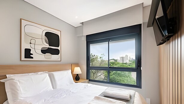 A hotel room with a bed, a nightstand, a lamp, a TV, and a large window with a city view. - Uwin 403 - Apartment 10 minutes from Chucri Zaidan (São Paulo)