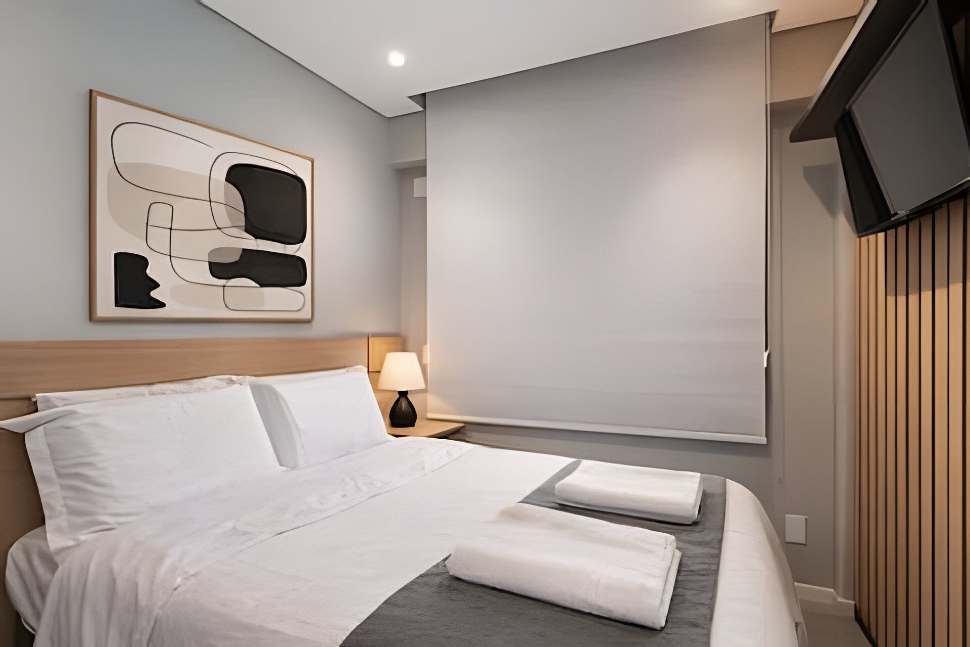 Quarto de hotel com cama, mesa de cabeceira com abajur, obra de arte abstrata emoldurada, televisão e janela com persianas.